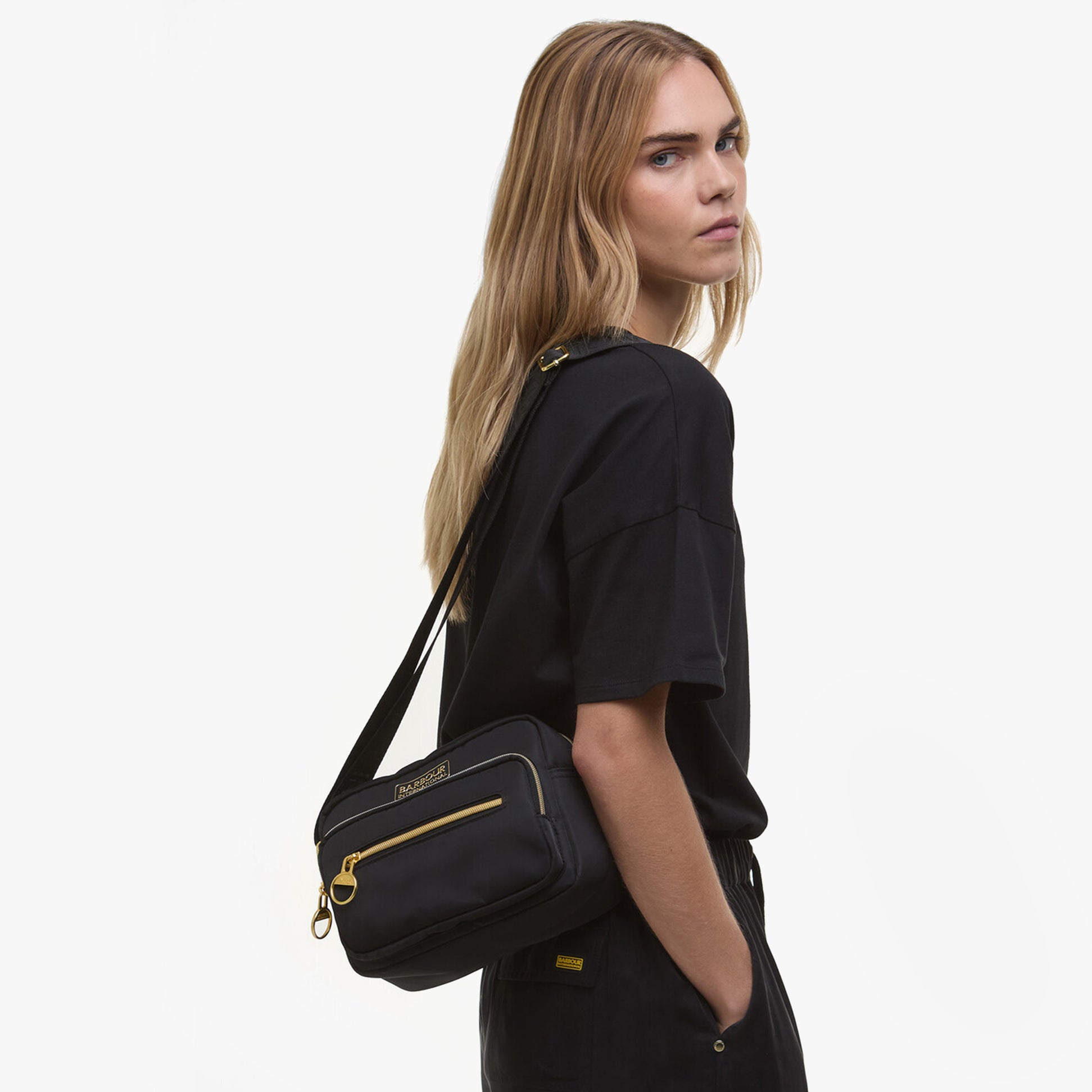 Barbour B.INTL QUALIFY Womens Crossbody Bag Black side view on model, showcasing adjustable strap and zipped pockets.