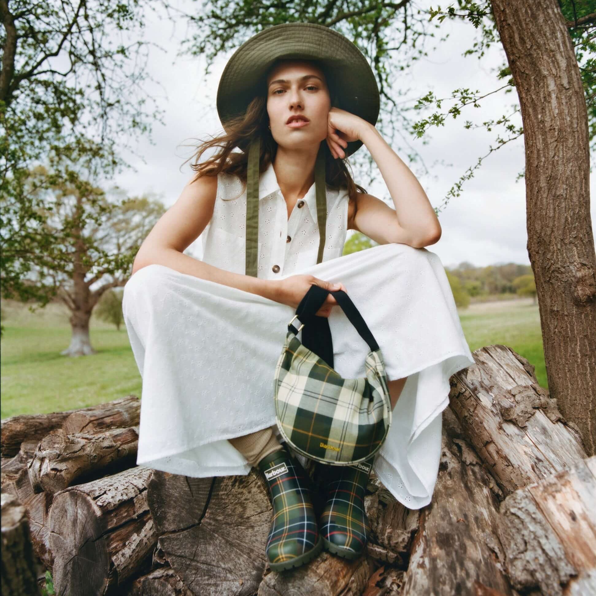 Barbour WILTON Womens Ankle Wellington Boots Classic Tartan worn by model sitting on a tree stump, side view, paired with plaid bag.