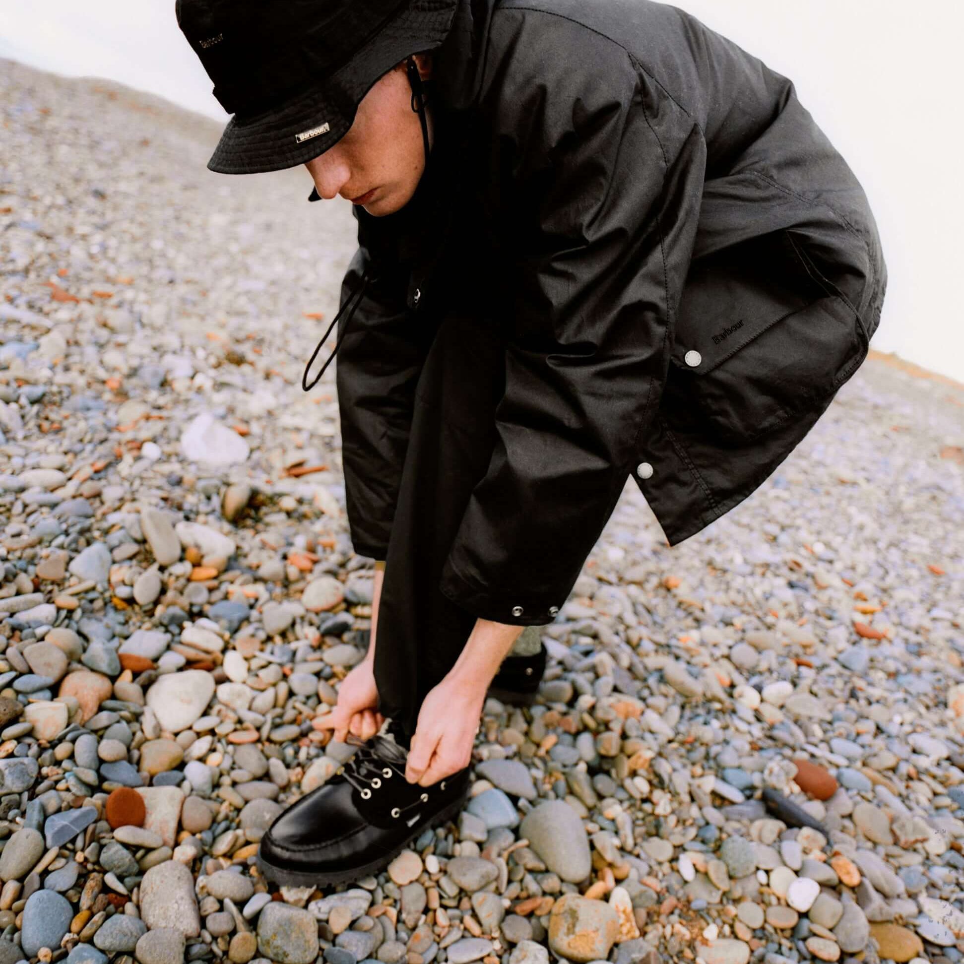 Barbour DECK Mens Boat Shoe Black worn by a person lacing up on a pebbly beach, showcasing the shoe's design.