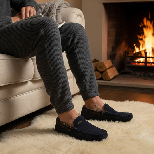Barbour MONTY Mens Slippers Navy worn by a man sitting on a sofa by a fireplace with cozy decor.