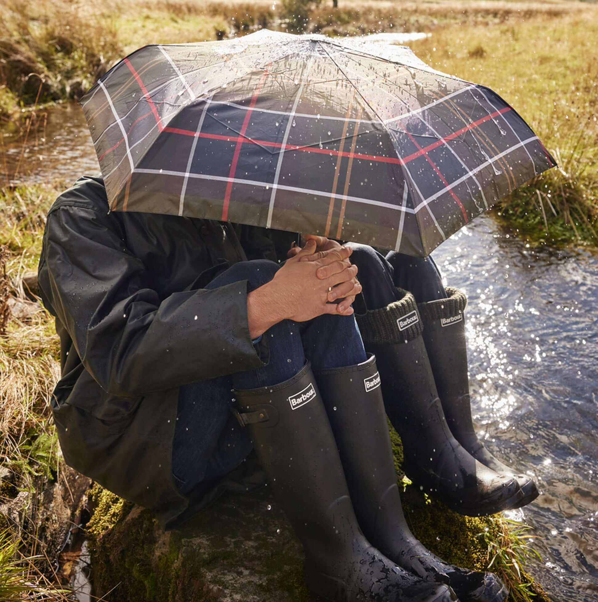 Barbour TARTAN FULL LENGTH Classic Umbrella shown in use under a rainy setting near a stream, providing stylish protection.