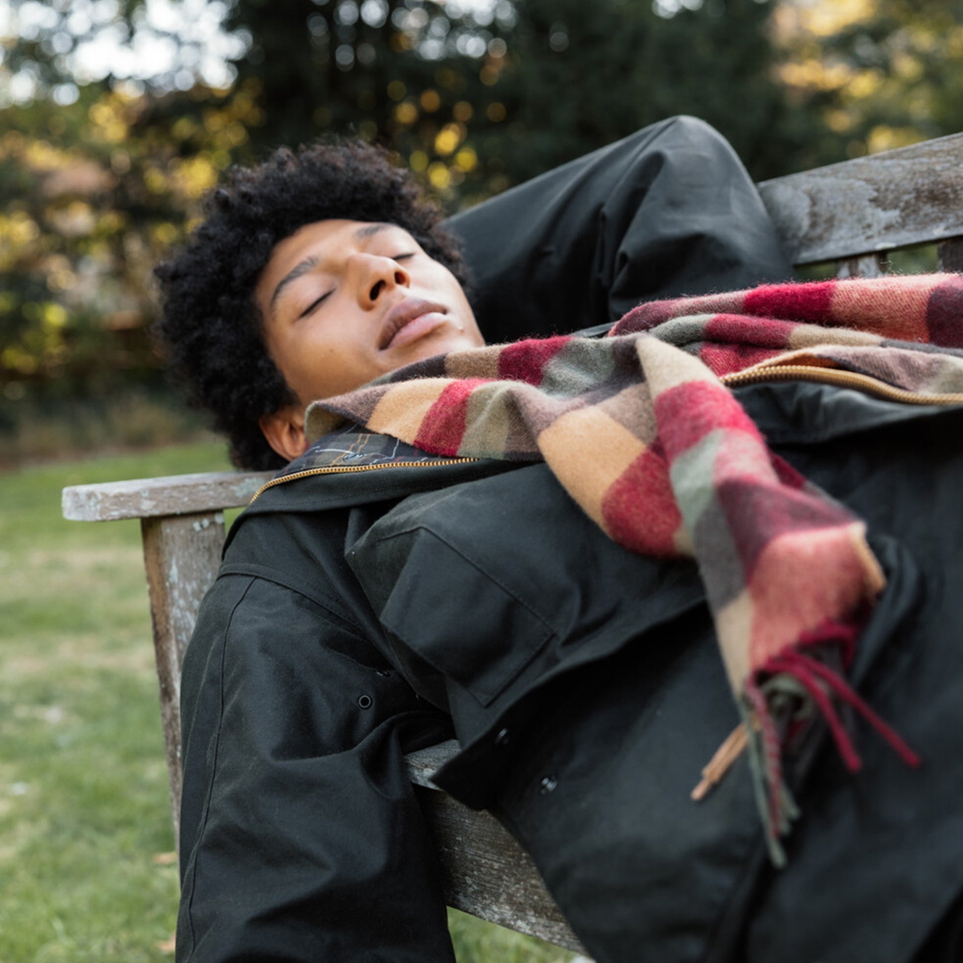 Barbour LAMBSWOOL TATTERSALL Scarf Unisex Adults Tawny Port detail view on model wearing black jacket, relaxing on bench outdoors.