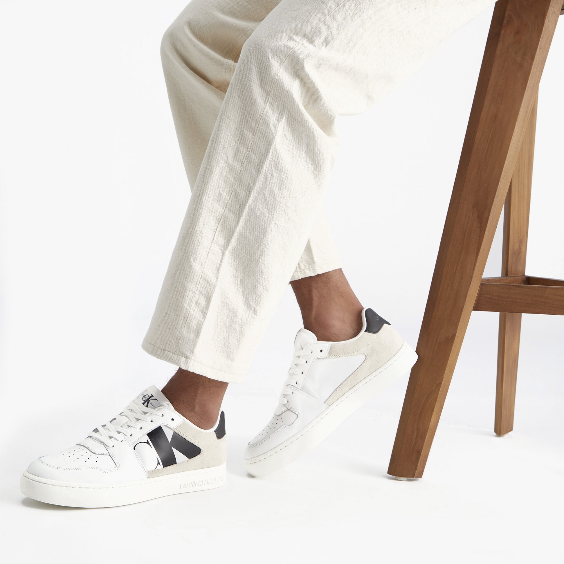 Calvin Klein CLASSIC CUPSOLE Mens Trainers Bright White/Black side view worn with casual outfit and stylish wooden chair.