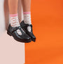 Hush Puppies FIONA Girls Leather School Shoes Black side view with pink socks on a model against an orange background.