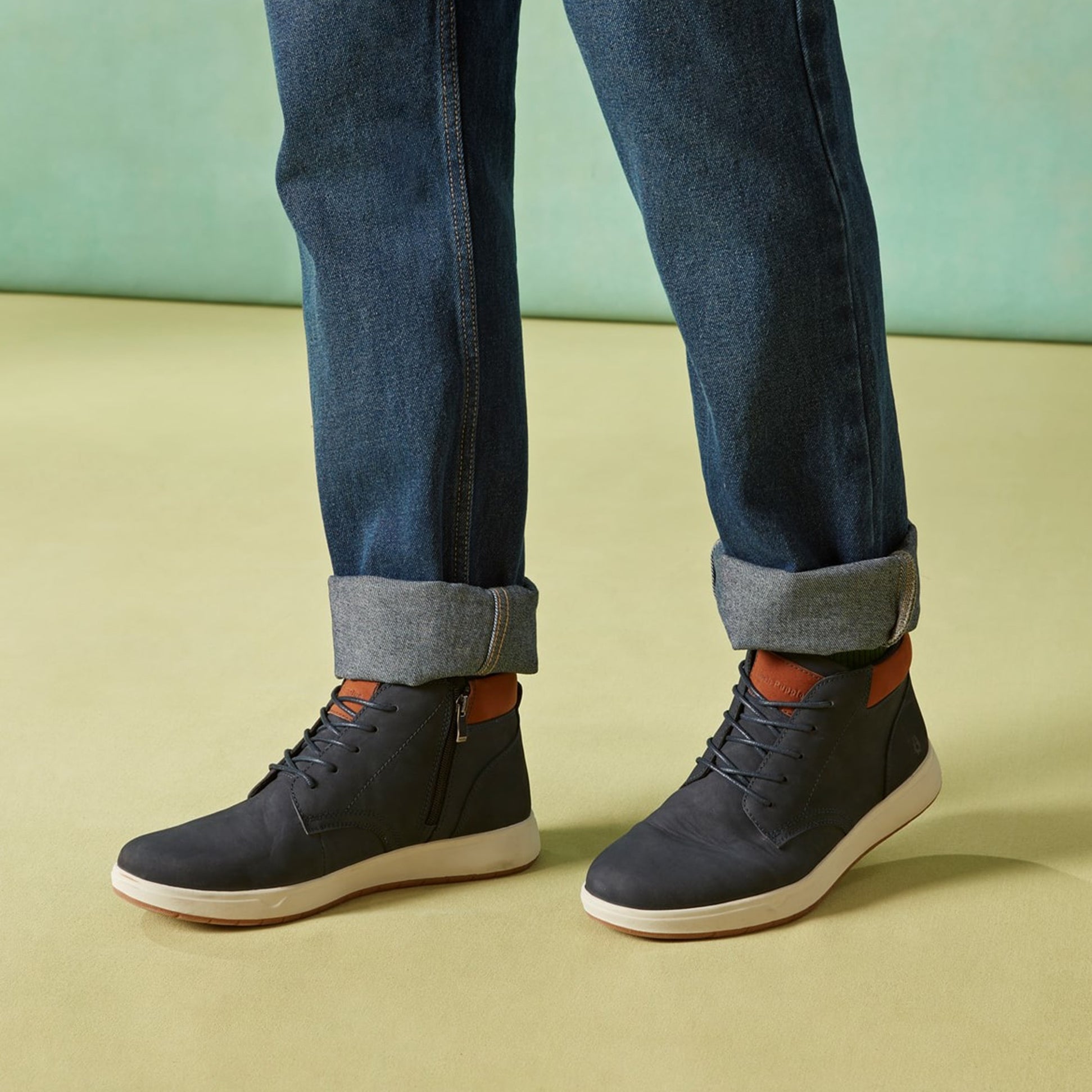 Hush Puppies BRONKO Mens Chukka Boots Navy side view worn with cuffed jeans on a yellow floor.