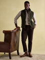 Joules GREENFIELD Mens Gilet Green side view on model, paired with a checked shirt and brown pants, standing near a leather chair.