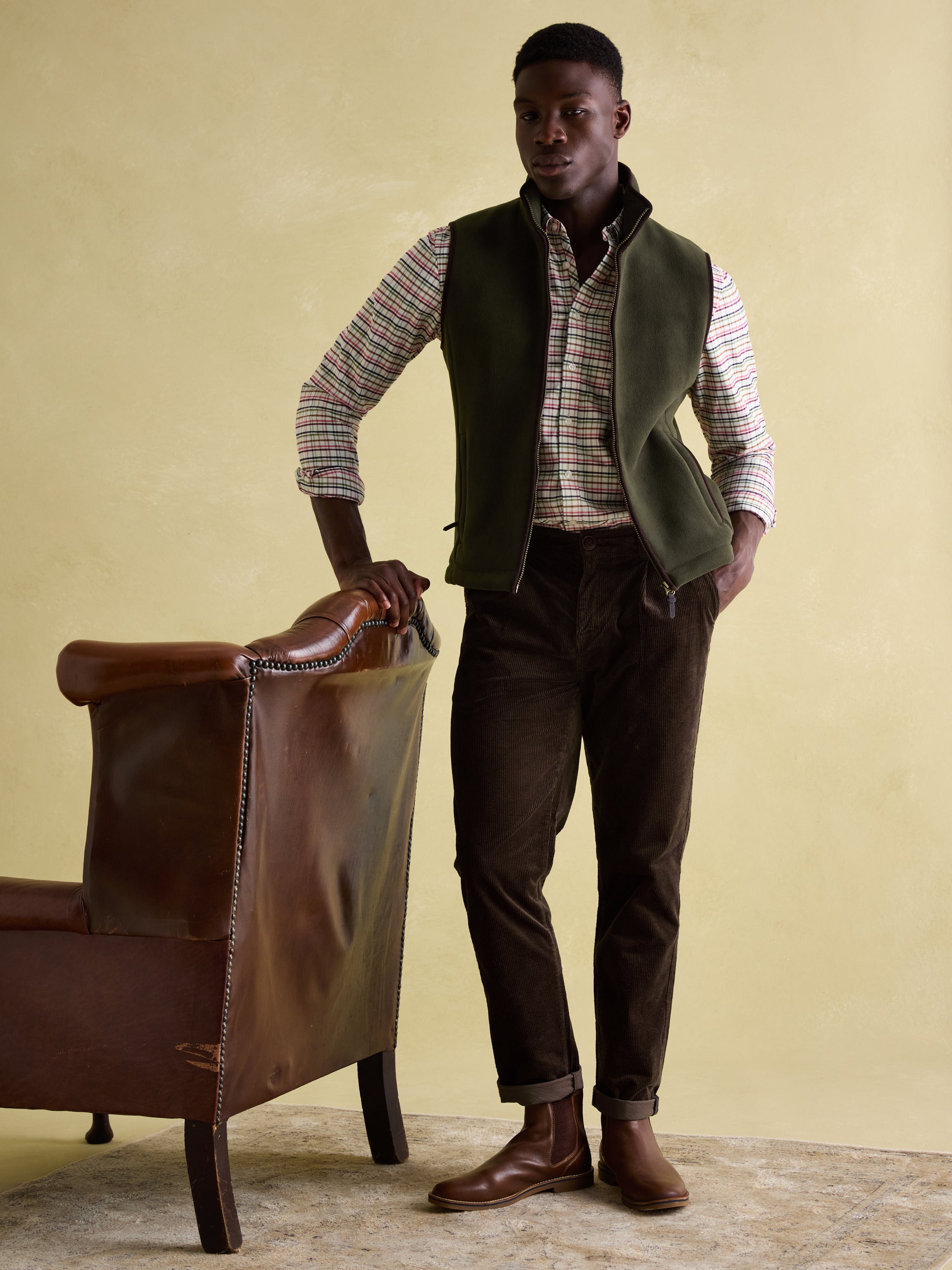 Joules GREENFIELD Mens Gilet Green side view on model, paired with a checked shirt and brown pants, standing near a leather chair.
