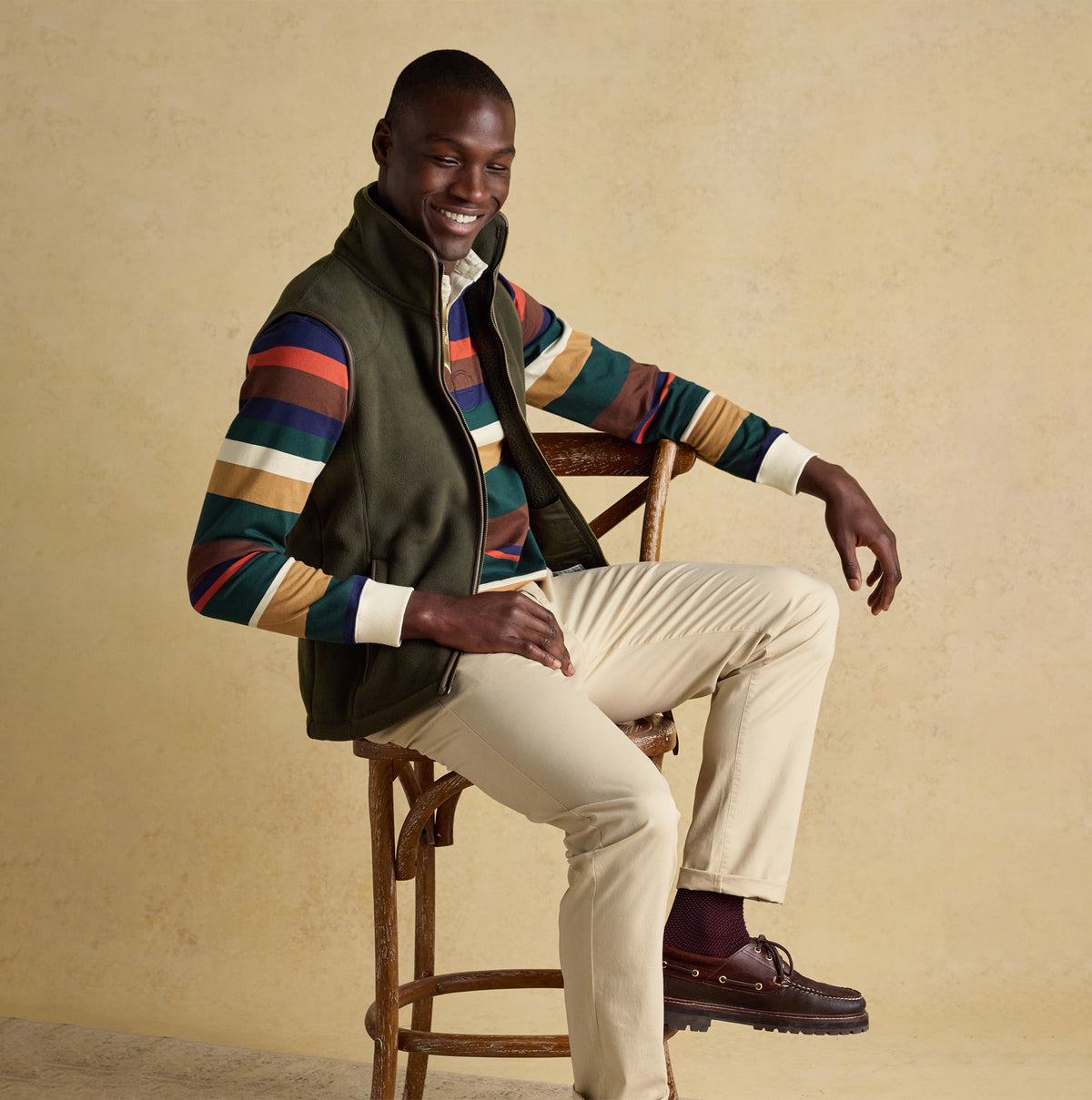 Joules HERITAGE FLEECE Mens Gilet Green front view on model, styled with colorful striped shirt and seated on chair.