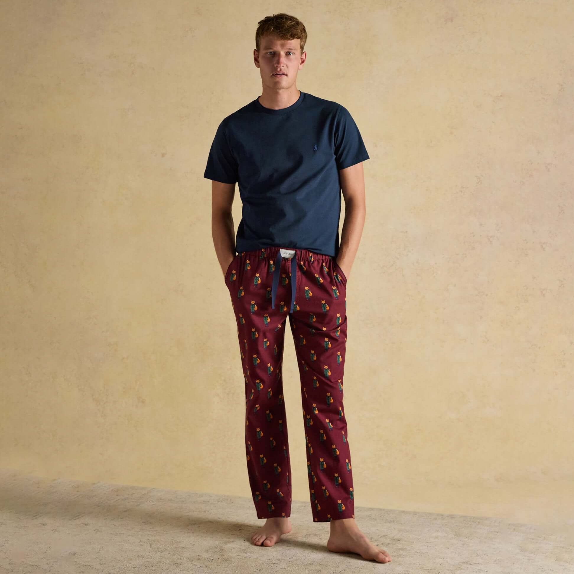 Joules DOZER Mens Pyjama Trousers Burgundy Red front view with a model wearing a navy top and burgundy patterned trousers.