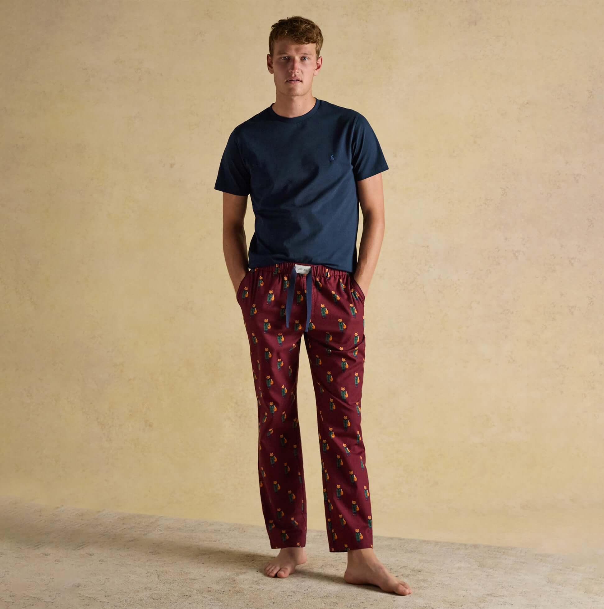 Joules DOZER Mens Pyjama Trousers Burgundy Red front view with a model wearing a navy top and burgundy patterned trousers.