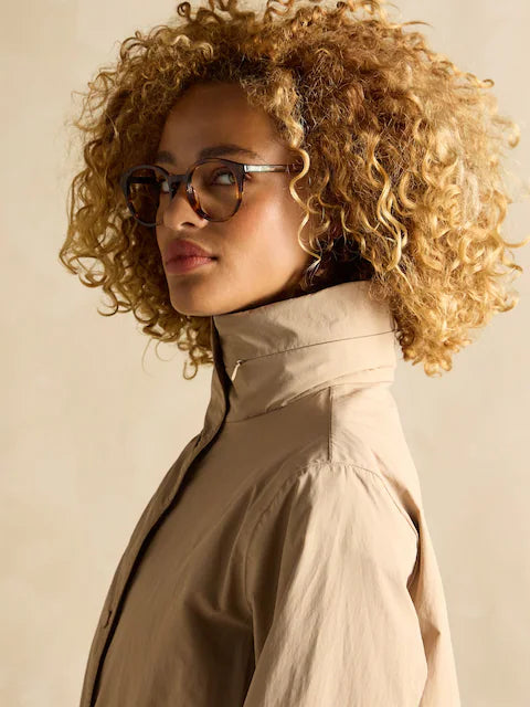 Joules Hensley Womens Waterproof Longline Coat with Packable Hood Tan Brown side view on model with curly hair and glasses.
