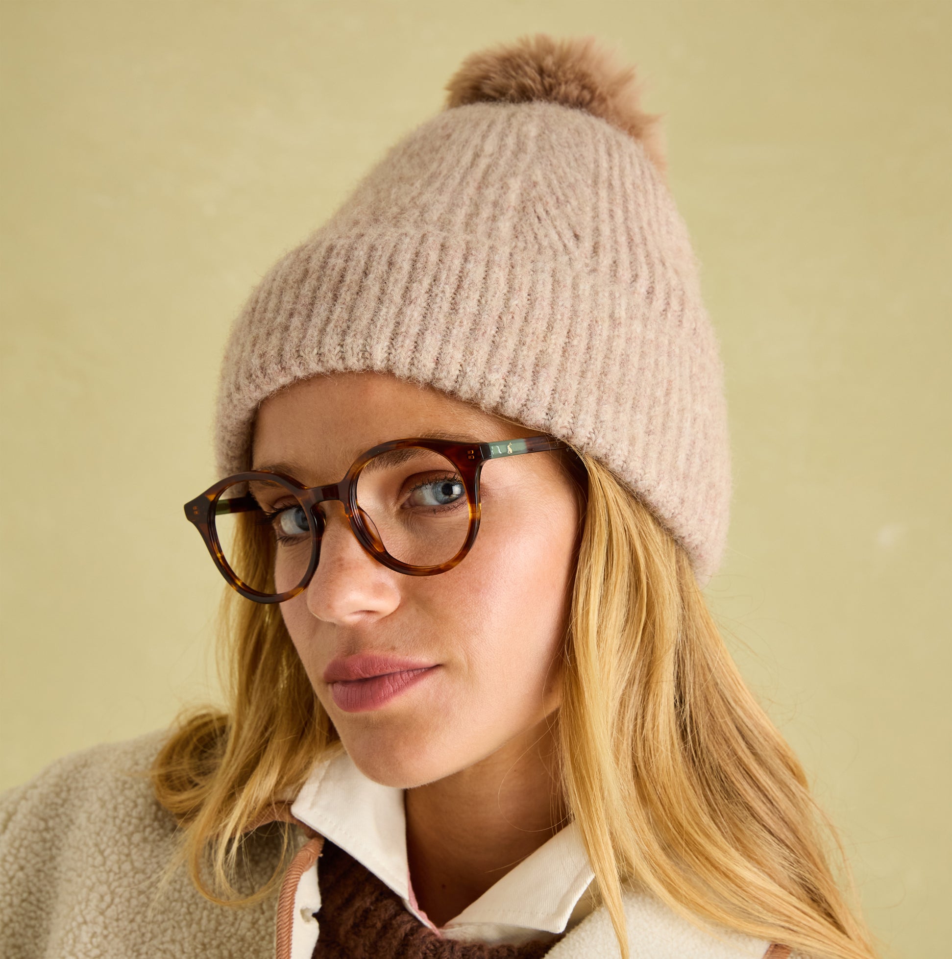 Joules EFFIE Womens Knitted Bobble Hat Camel worn by a model with glasses, close-up detail view showcasing the hat's texture.