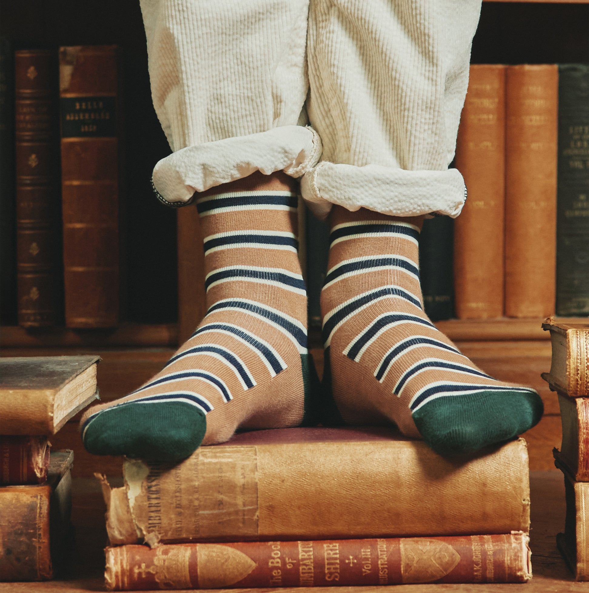 Joules KINGSBURY 2-Pack Mens Socks Navy Camel knee-level view showing striped design on feet with books in background.