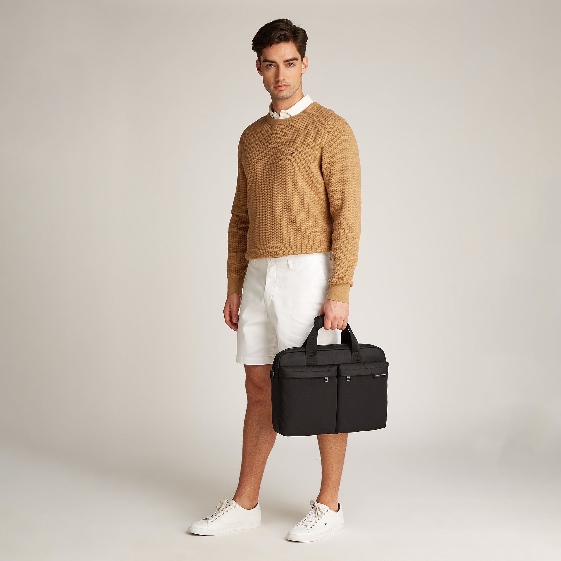 Tommy Hilfiger Rubberised Logo Laptop Bag in black, front view held by a man in a tan sweater and white shorts.