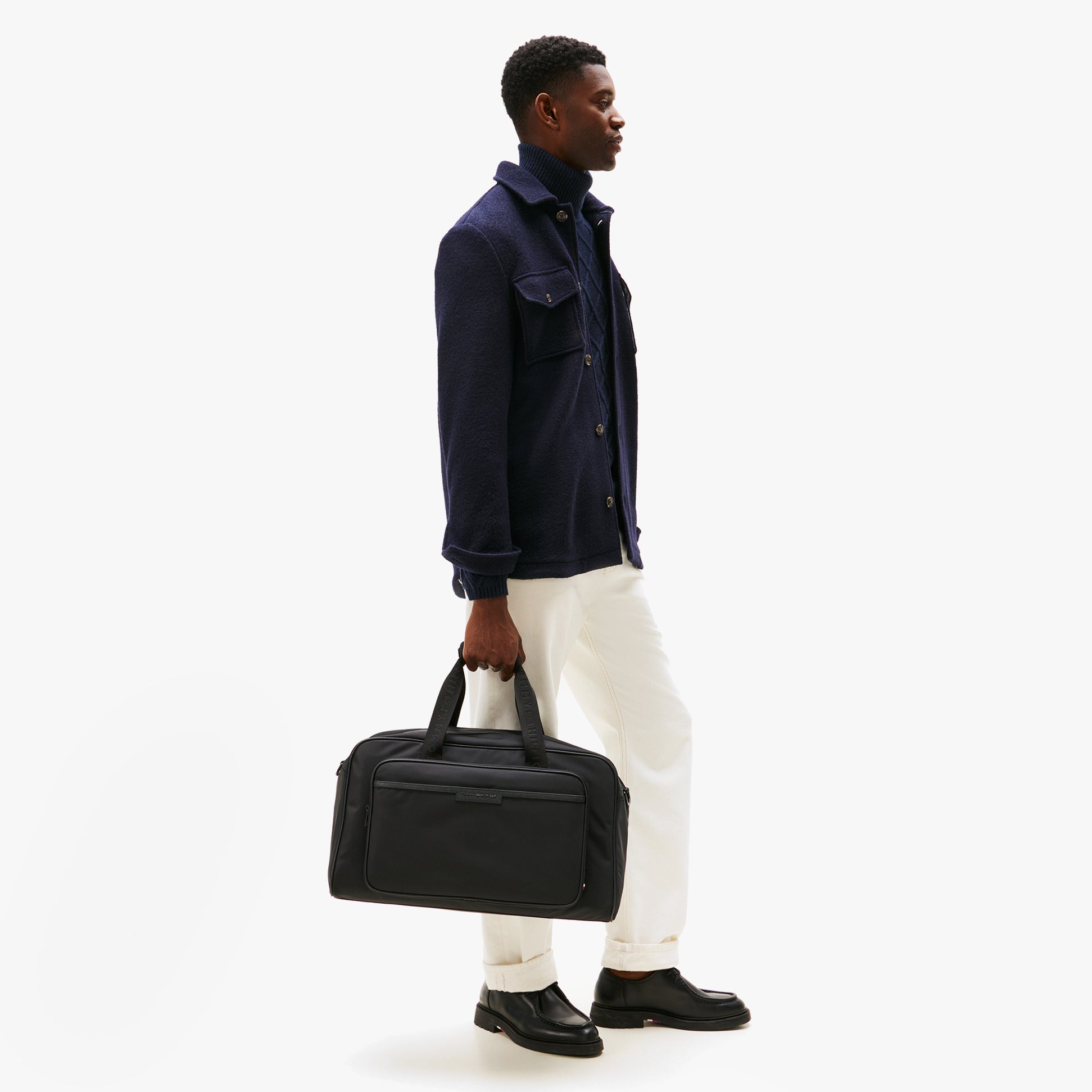 Tommy Hilfiger REPREVE DUFFLE Mens Duffle Bag Black side view on model, stylishly held in hand, casual outfit, urban setting.