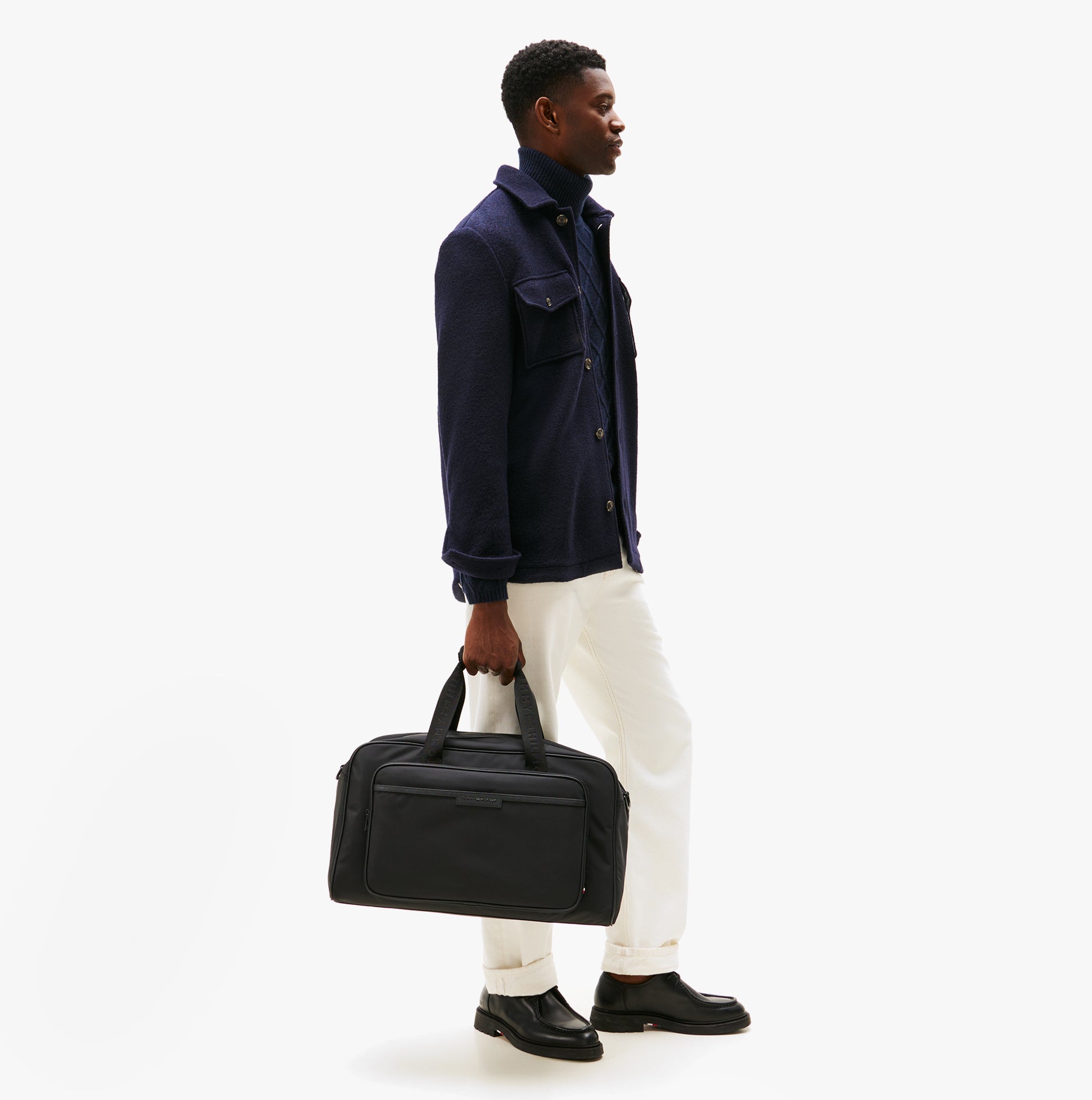 Tommy Hilfiger REPREVE DUFFLE Mens Duffle Bag Black side view on model, stylishly held in hand, casual outfit, urban setting.