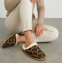 Totes Isotoner ANIMAL FUR Womens Mule Slippers Animal Print side view displayed on a model sitting with light-colored pants.