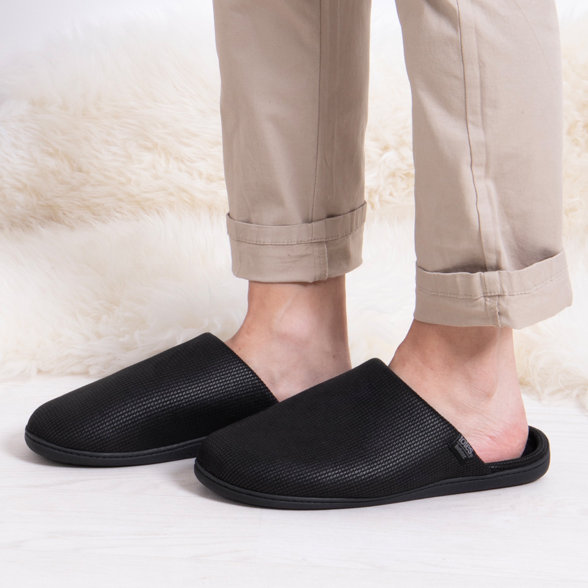 Totes Isotoner SQUARE PATTERNED SMART MULE Mens Mule Slippers Black side view on feet with beige pants.
